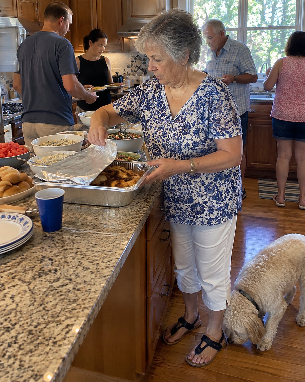 At a picnic table at a family reunion mid-morning, a woman reaches for a foil pan while a dog noses around beneath her feet and relatives pass dishes behind her.