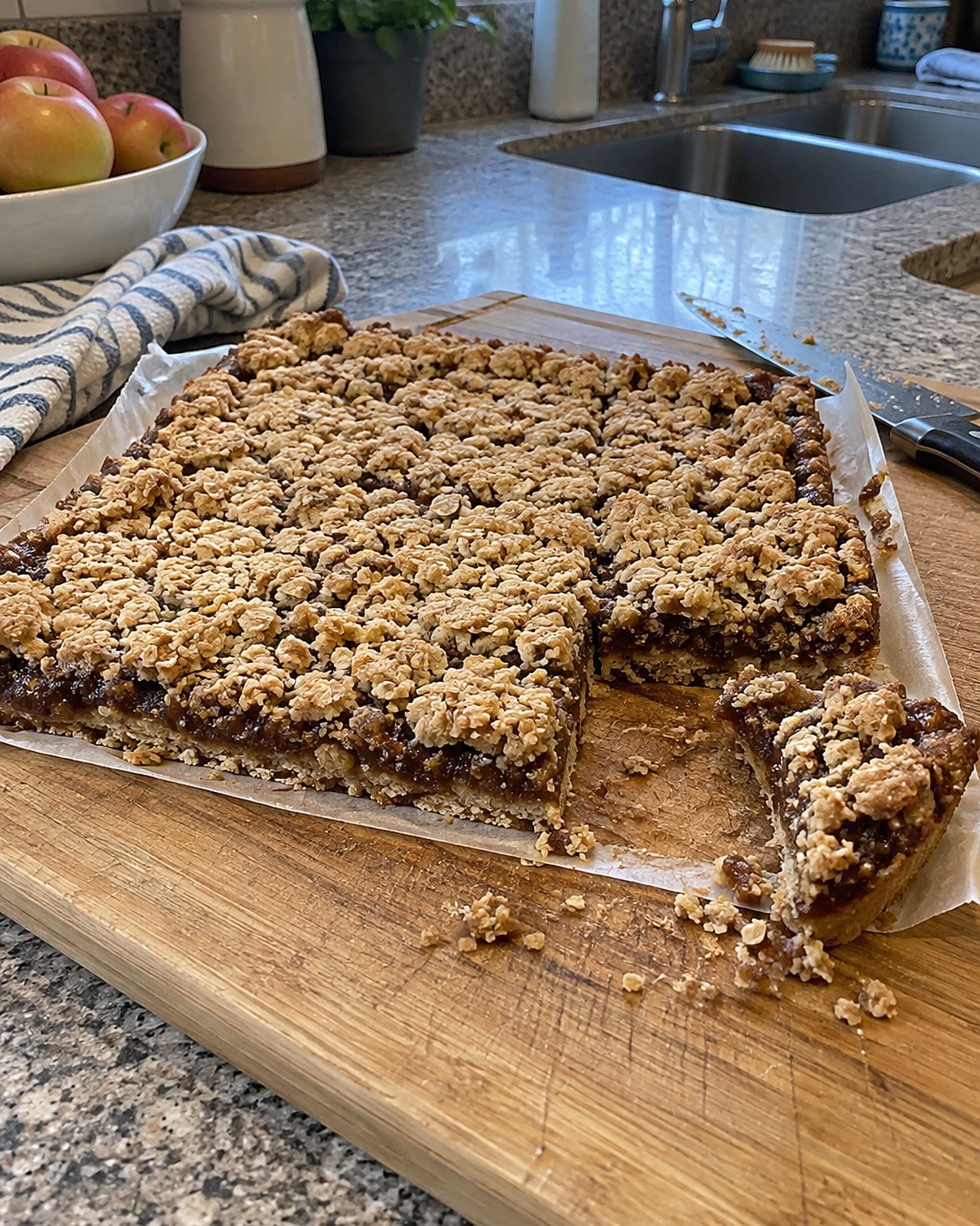 A slab of date bars lifted by parchment handles onto a wooden cutting board.
