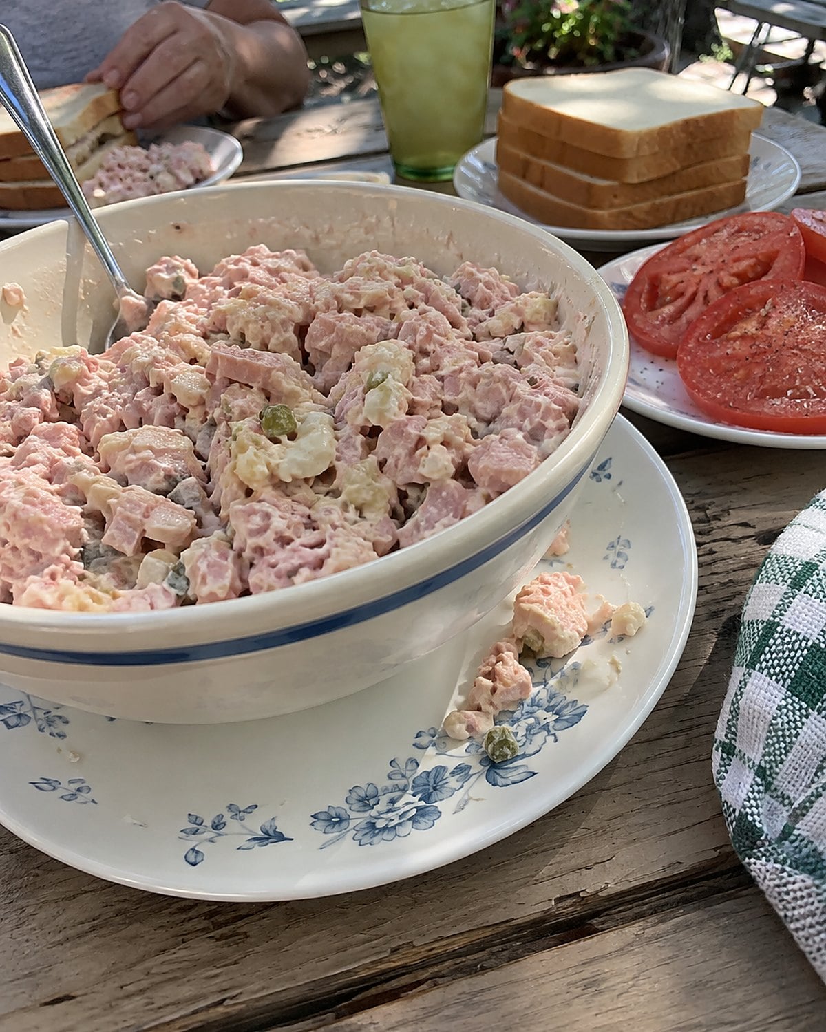 A bowl of bologna salad beside sliced tomatoes and a stack of bread on a picnic table.