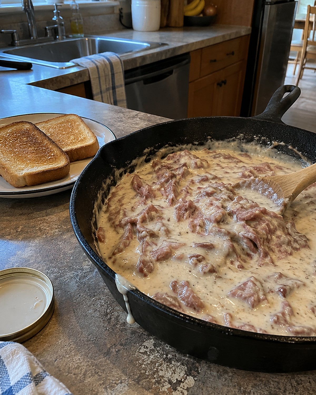 A cast-iron skillet of creamed dried beef beside two slices of toast on a worn counter.