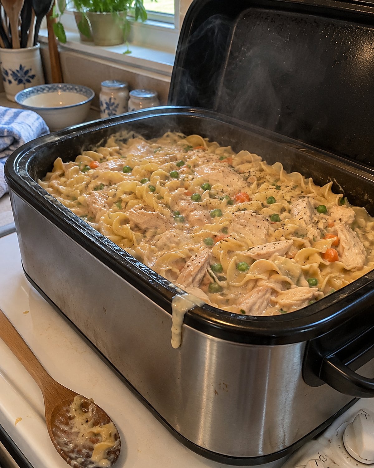 An electric roaster of chicken and noodles on a white stove, its lid tilted against the wall.
