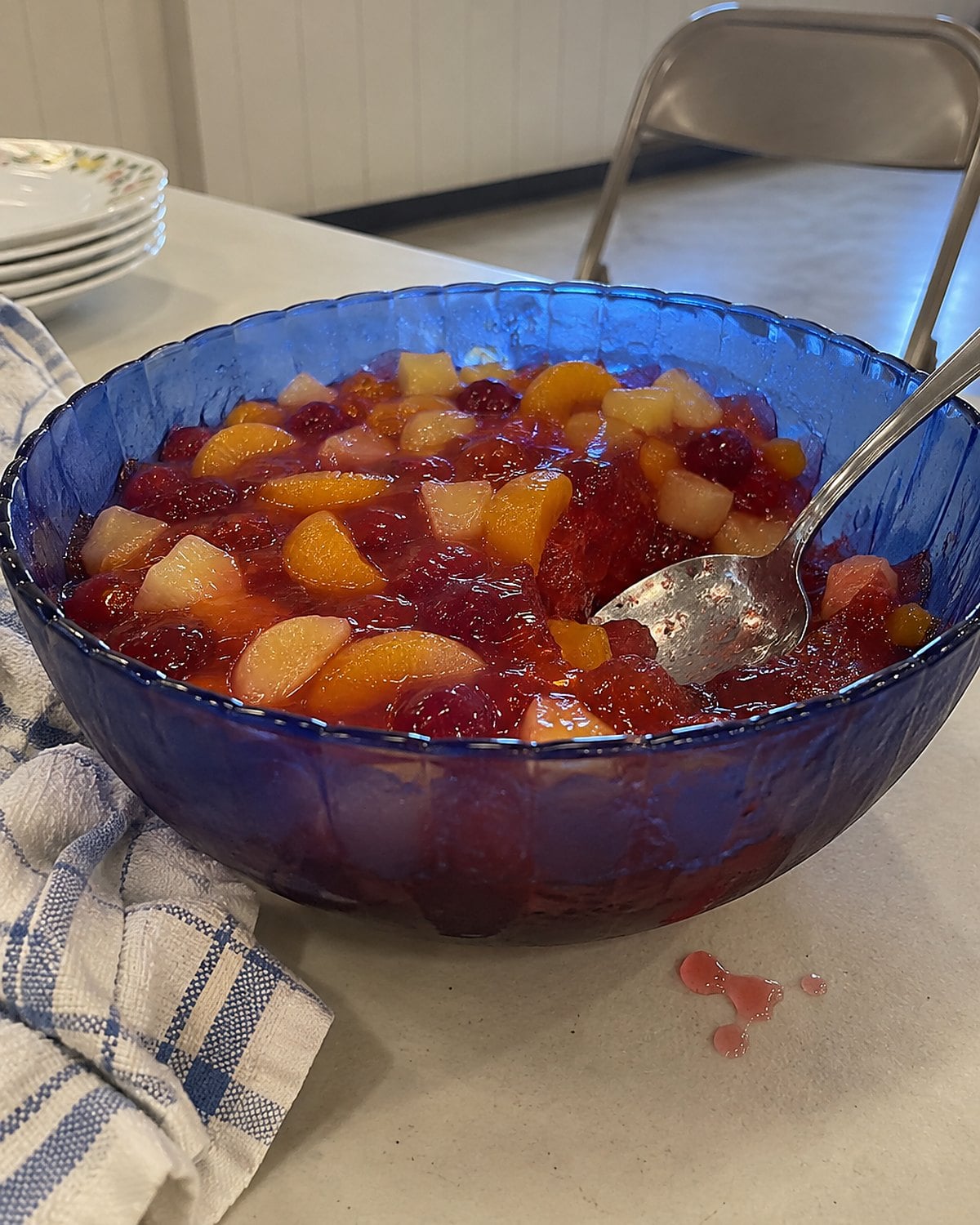 A blue bowl of fruit-filled gelatin on a church hall table, with a serving spoon resting against its rim.