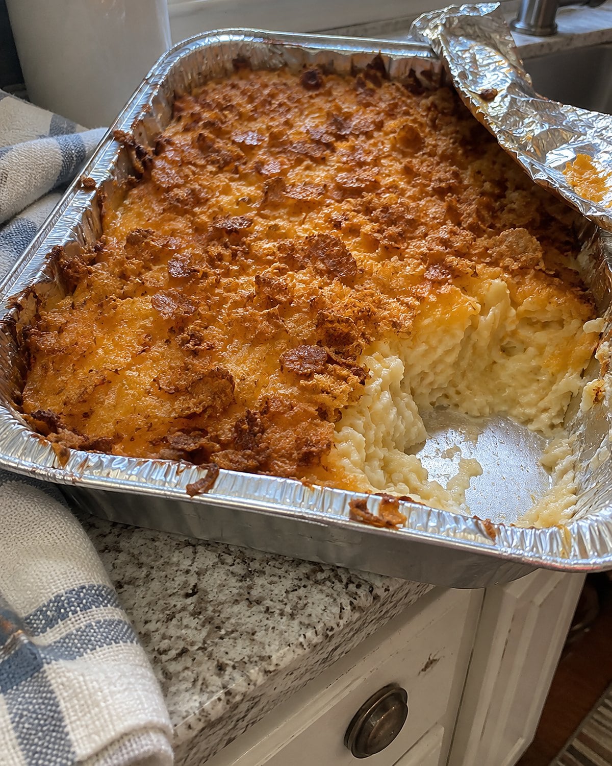 A foil pan of funeral potatoes on a kitchen counter, with one browned corner scooped out and the foil folded back.