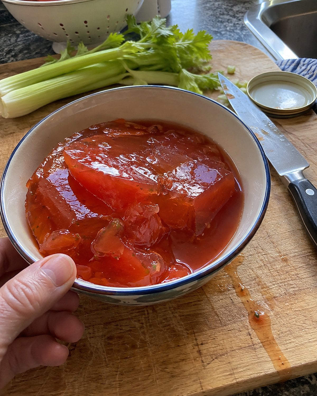 A small bowl of tomato aspic on a cutting board beside a knife and celery stalks.