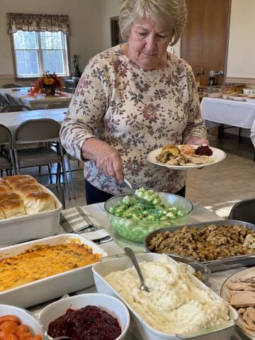 At a church-hall folding table during a mid-afternoon Thanksgiving potluck, a woman in her sixties stands alone spooning lime gelatin salad onto her plate, with open casserole dishes nearby.