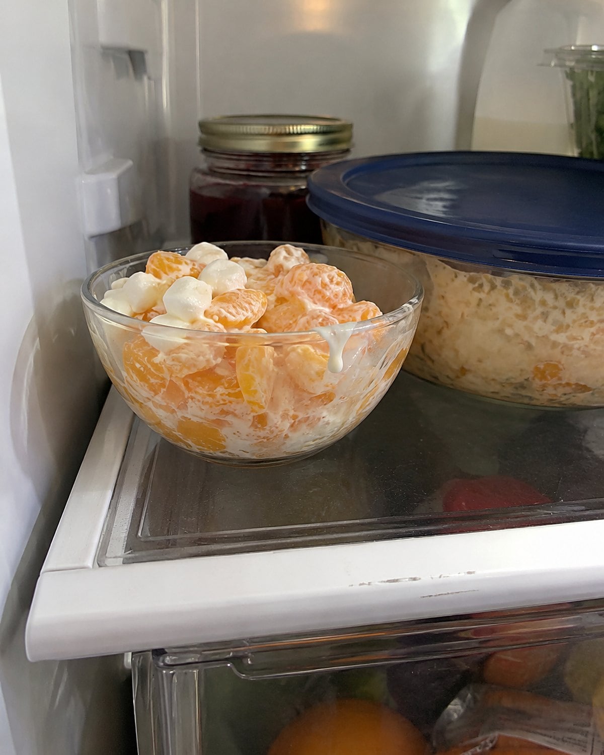 A small bowl of orange ambrosia sits on a refrigerator shelf beside a covered larger bowl.