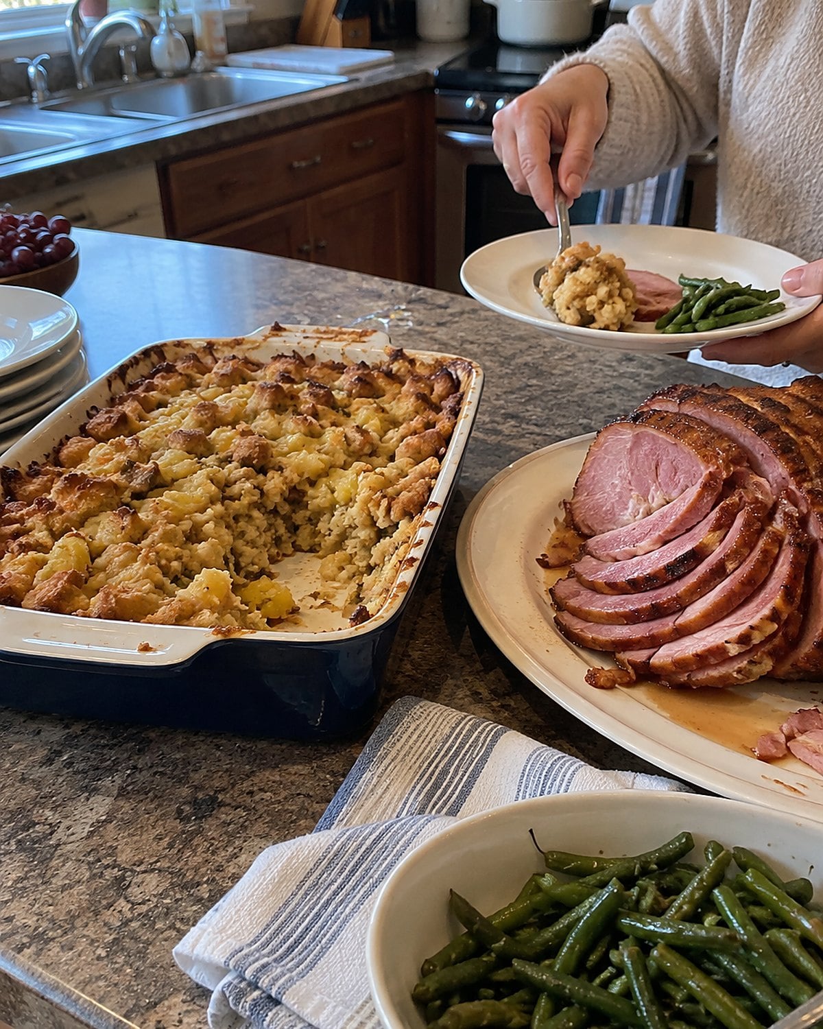 A corner scoop is missing from pineapple stuffing in a small baking dish beside a platter of ham.