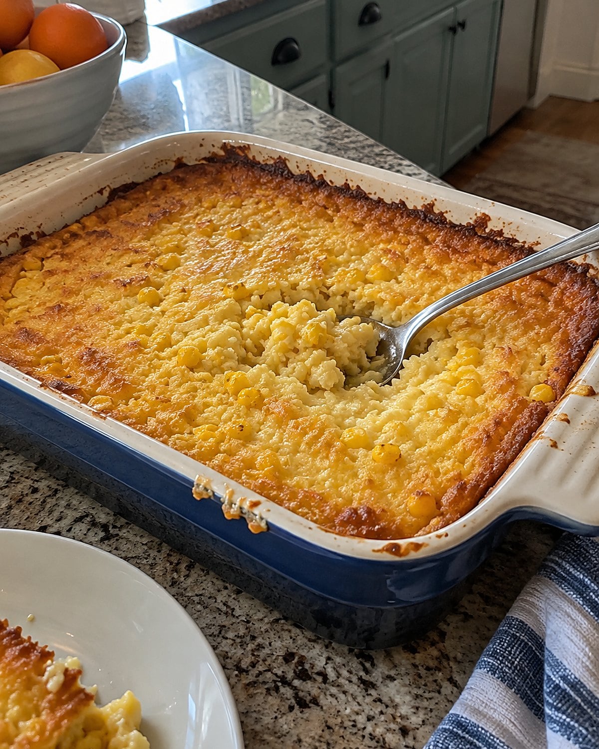 A spoon rests at the soft center of corn pudding in a baking dish with browned edges.