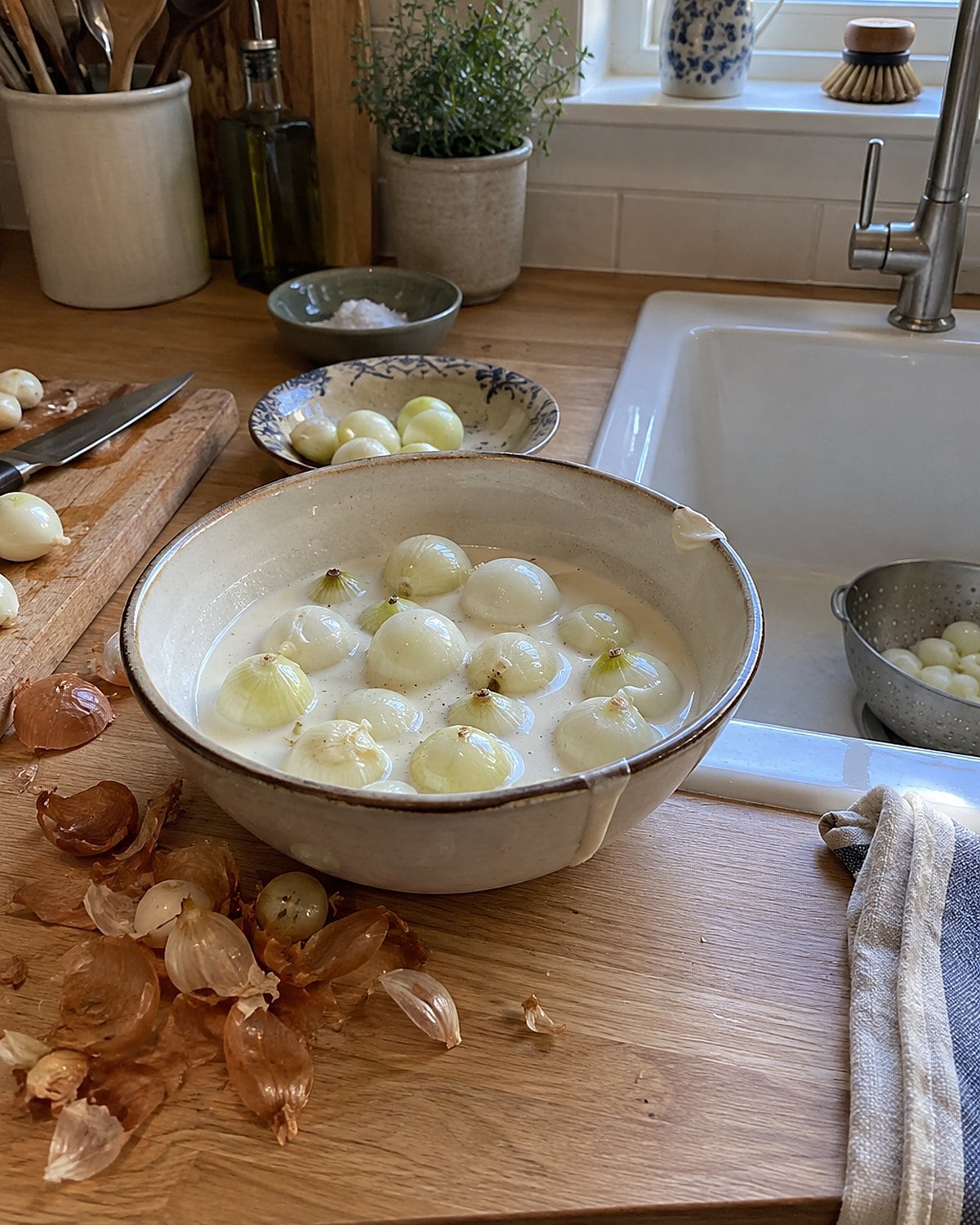 Peeled pearl onions in cream fill a shallow bowl beside onion skins at the sink.