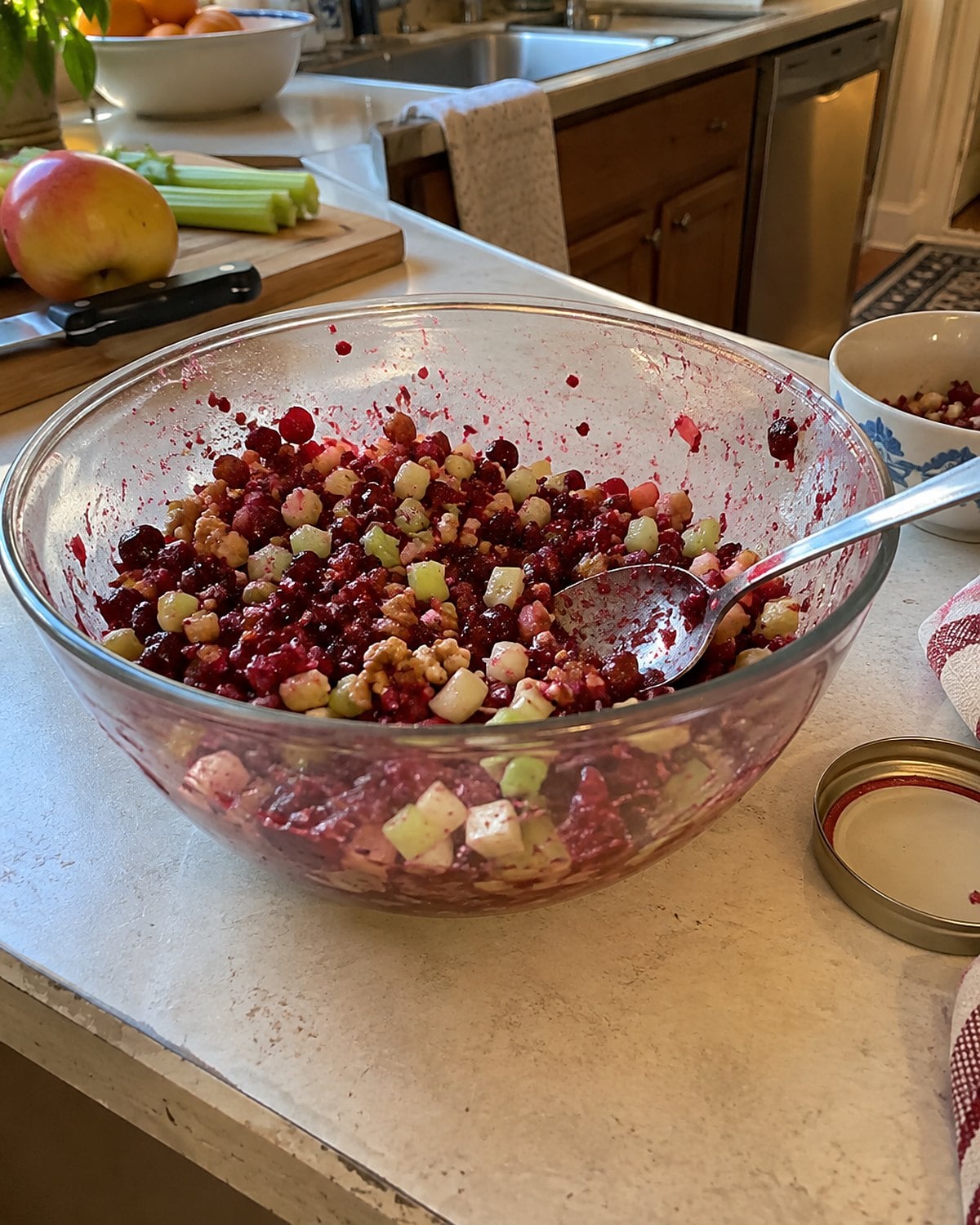 A glass bowl of chopped cranberry and celery salad sits beside a stained serving spoon on a counter.