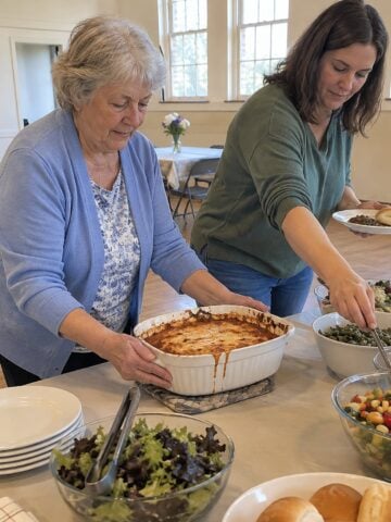 At a church-hall folding table mid-morning, a woman steadies a casserole dish while her daughter-in-law reaches past her for a serving spoon, neither looking at the camera.