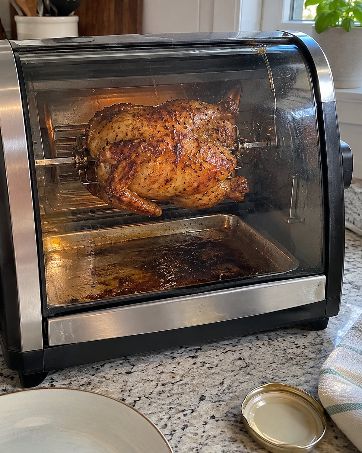 A countertop rotisserie with a browned chicken turning above its drip pan.