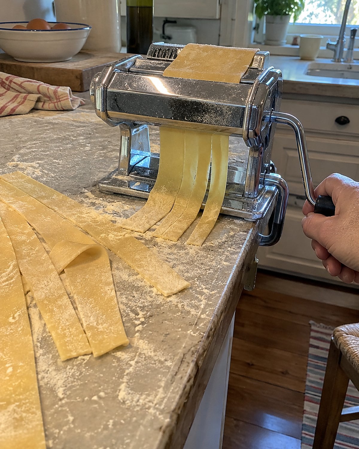 A hand-cranked pasta roller clamped to a counter beside uneven ribbons of dough.