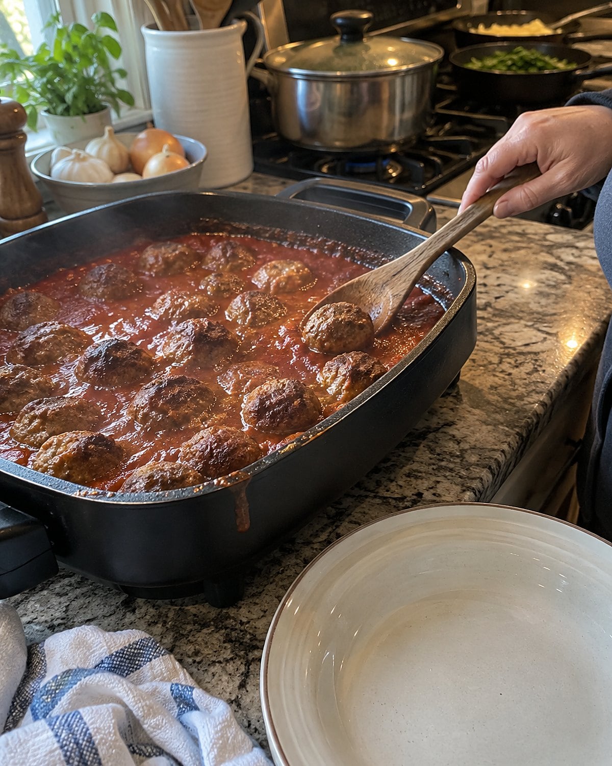 An electric skillet of meatballs on a kitchen counter beside a crowded stove.