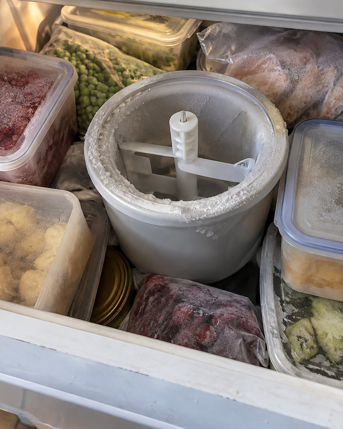 An ice cream maker bowl tucked among frozen food in a crowded freezer drawer.