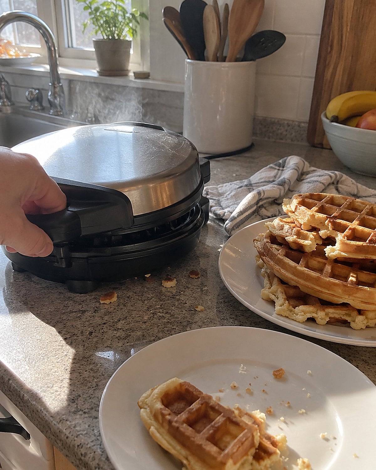 A waffle iron steaming on a counter beside an uneven stack of waffles.