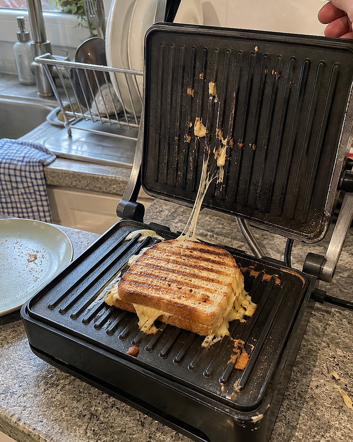 A sandwich press open at the sink, with melted cheese caught near its hinge.