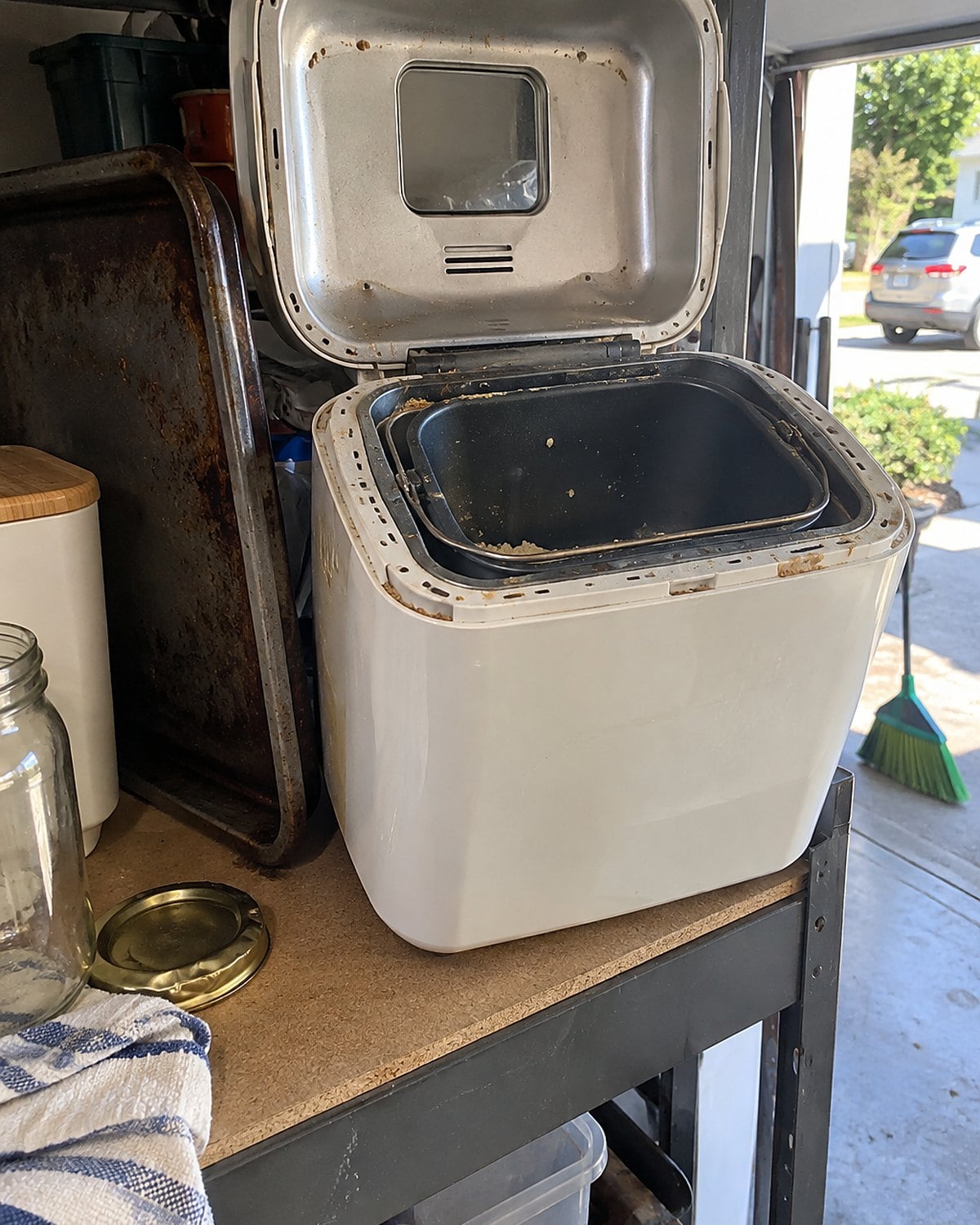 A bread machine with its lid open on a garage shelf beside a roasting pan.