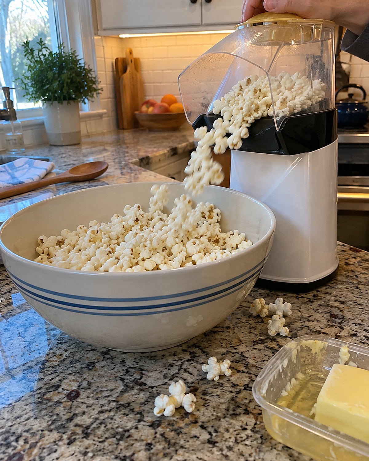 A hot-air popcorn popper aimed into a deep bowl, with its butter tray beside it.