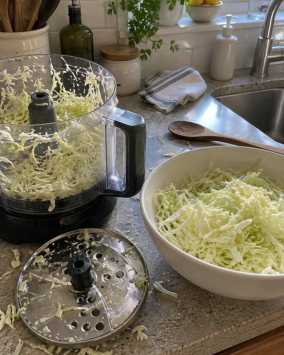 A food processor with its shredding disc beside a bowl of cabbage near the sink.