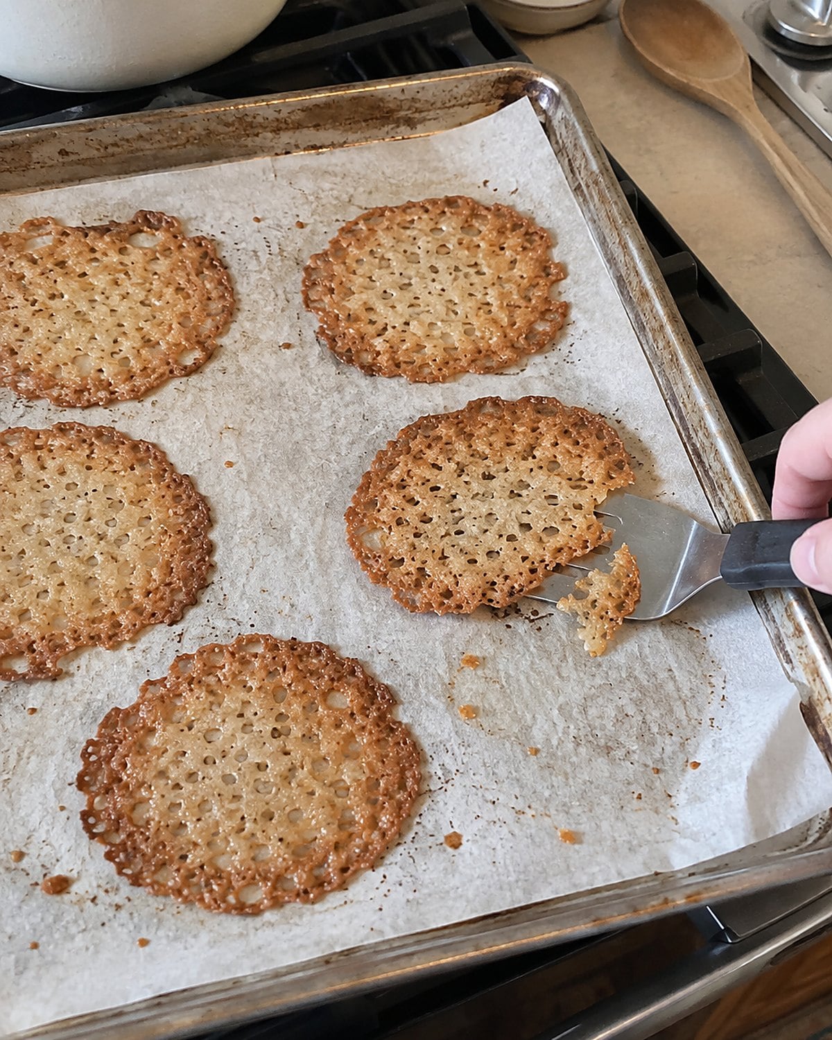 A thin spatula lifts one brown-edged lace cookie from a baking sheet on the stove.