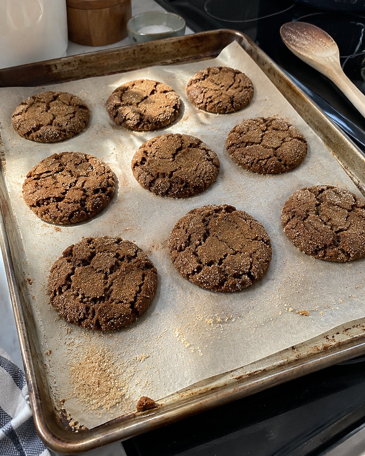 Cracked molasses cookies cool on a baking sheet with loose sugar near the corner.