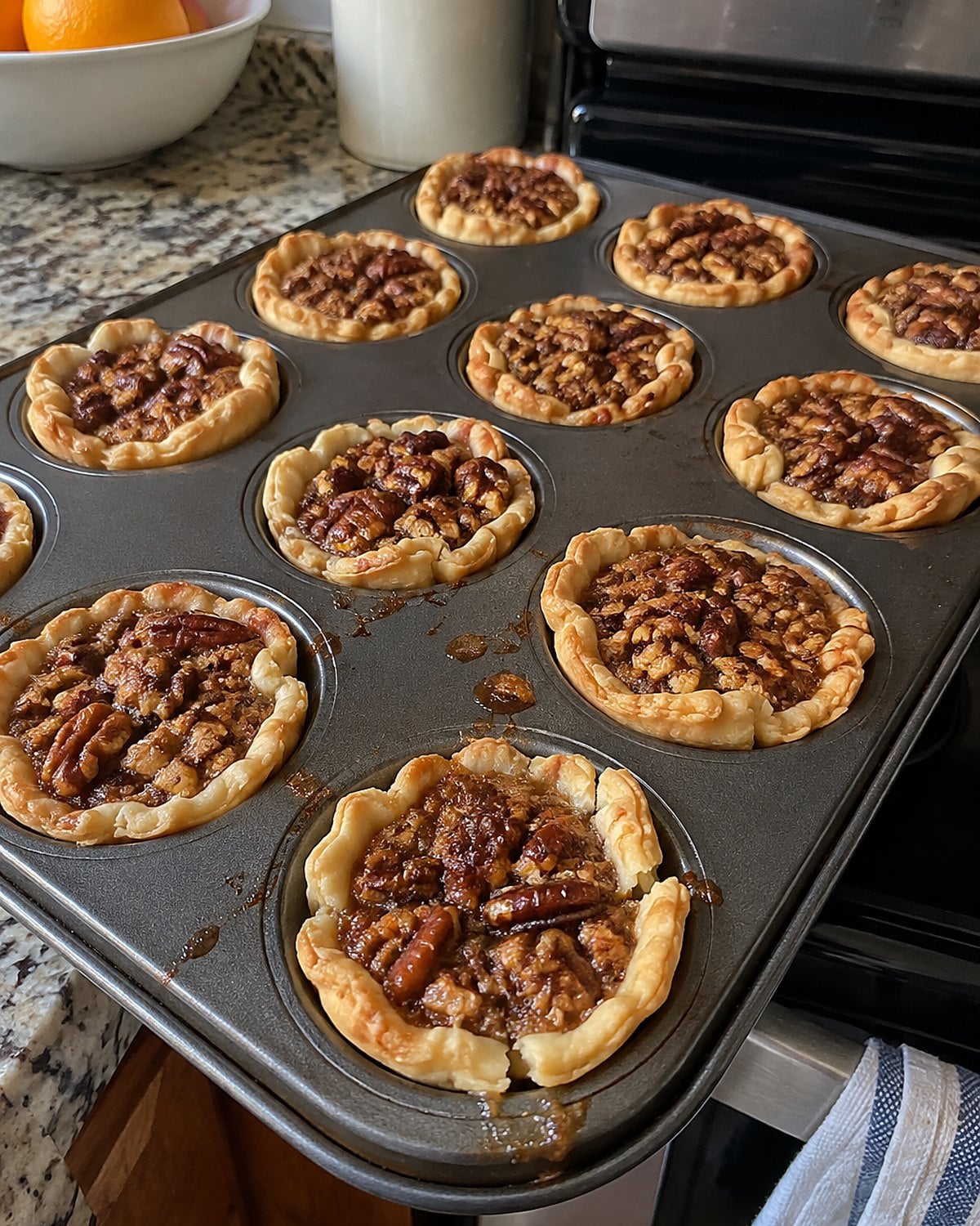 A mini muffin pan holds pecan tassies with one cracked pastry rim near the oven.