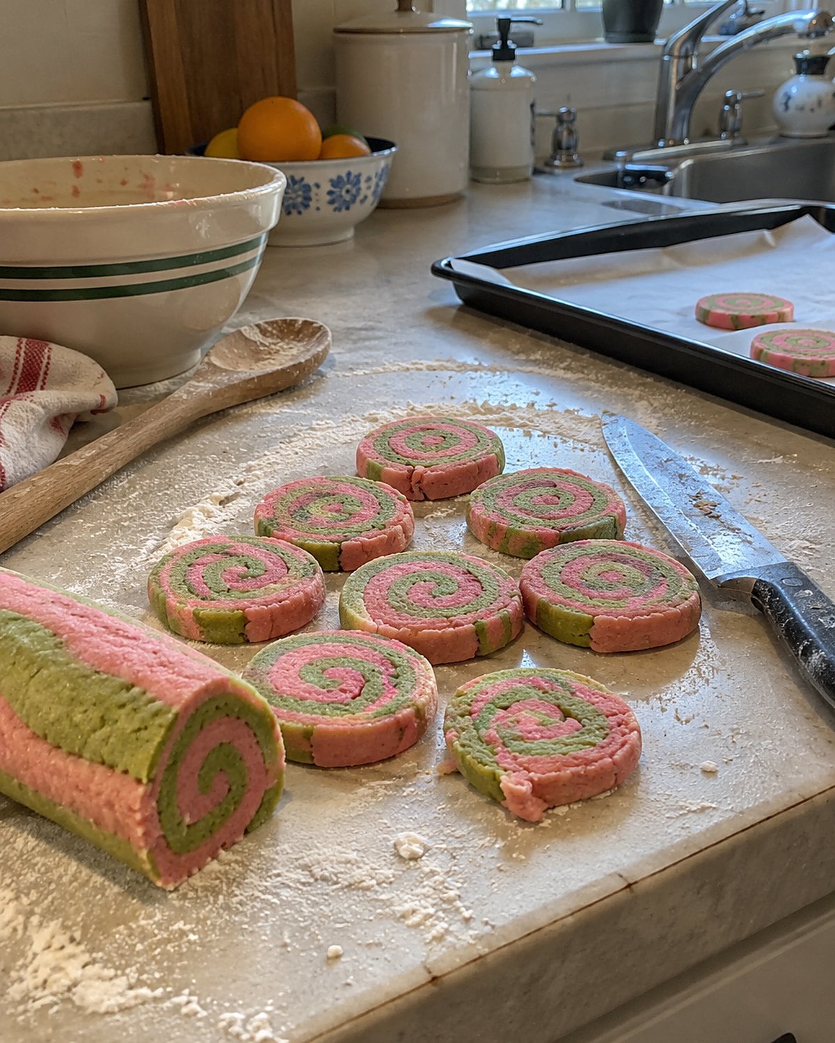 Slices of pink and green ribbon cookie dough lie beside a floured knife on a kitchen counter.