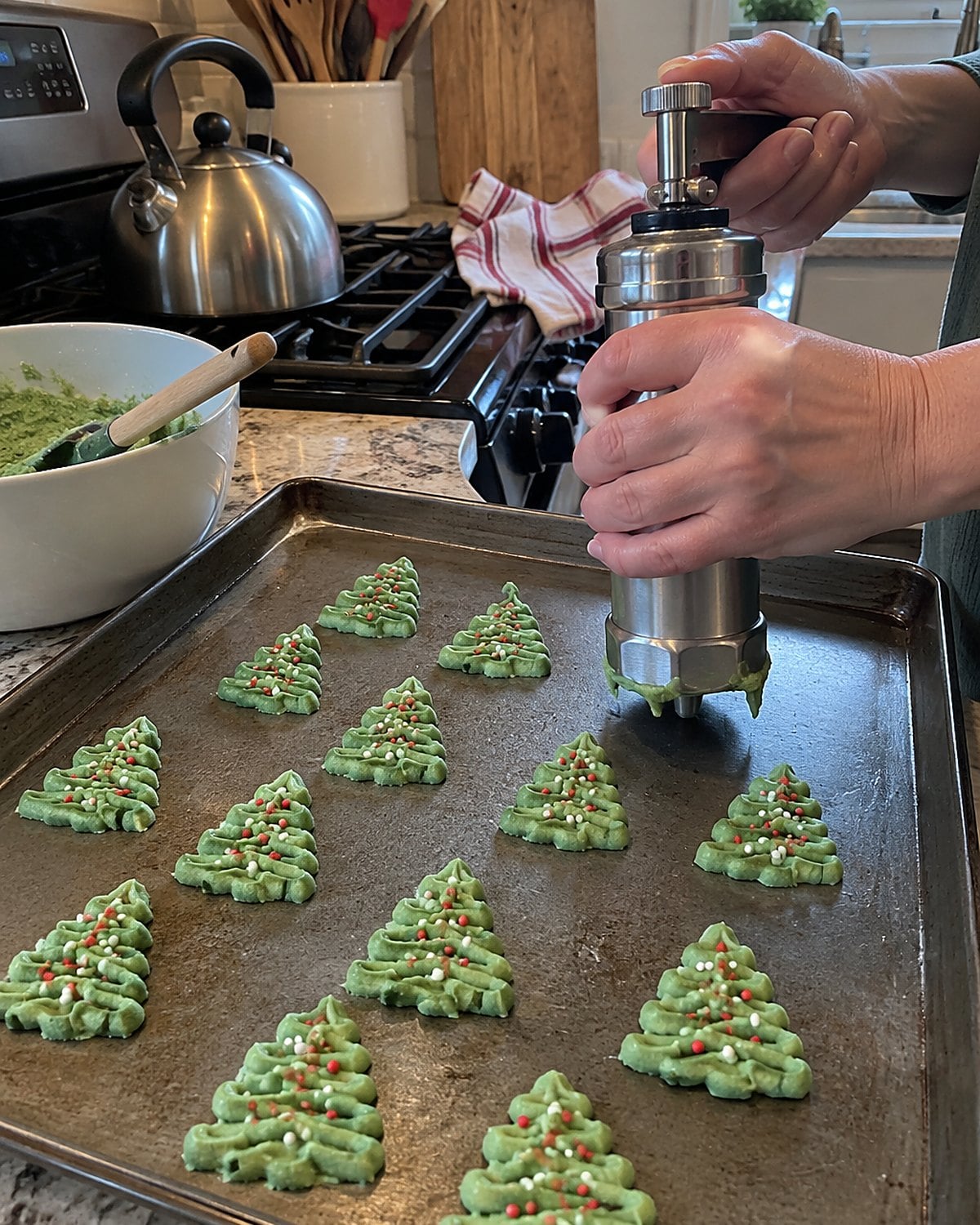 A metal cookie press and uneven spritz trees sit on an ungreased baking sheet beside the stove.