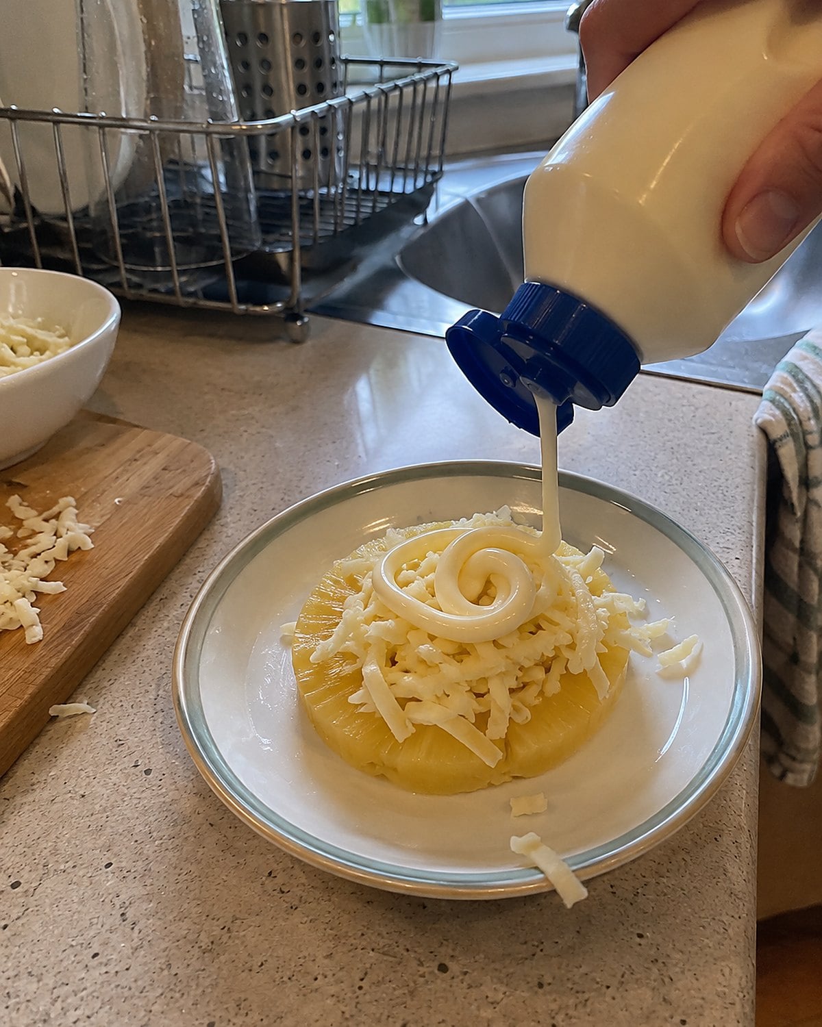 A pineapple ring with shredded cheese and mayonnaise on a small plate near the sink.