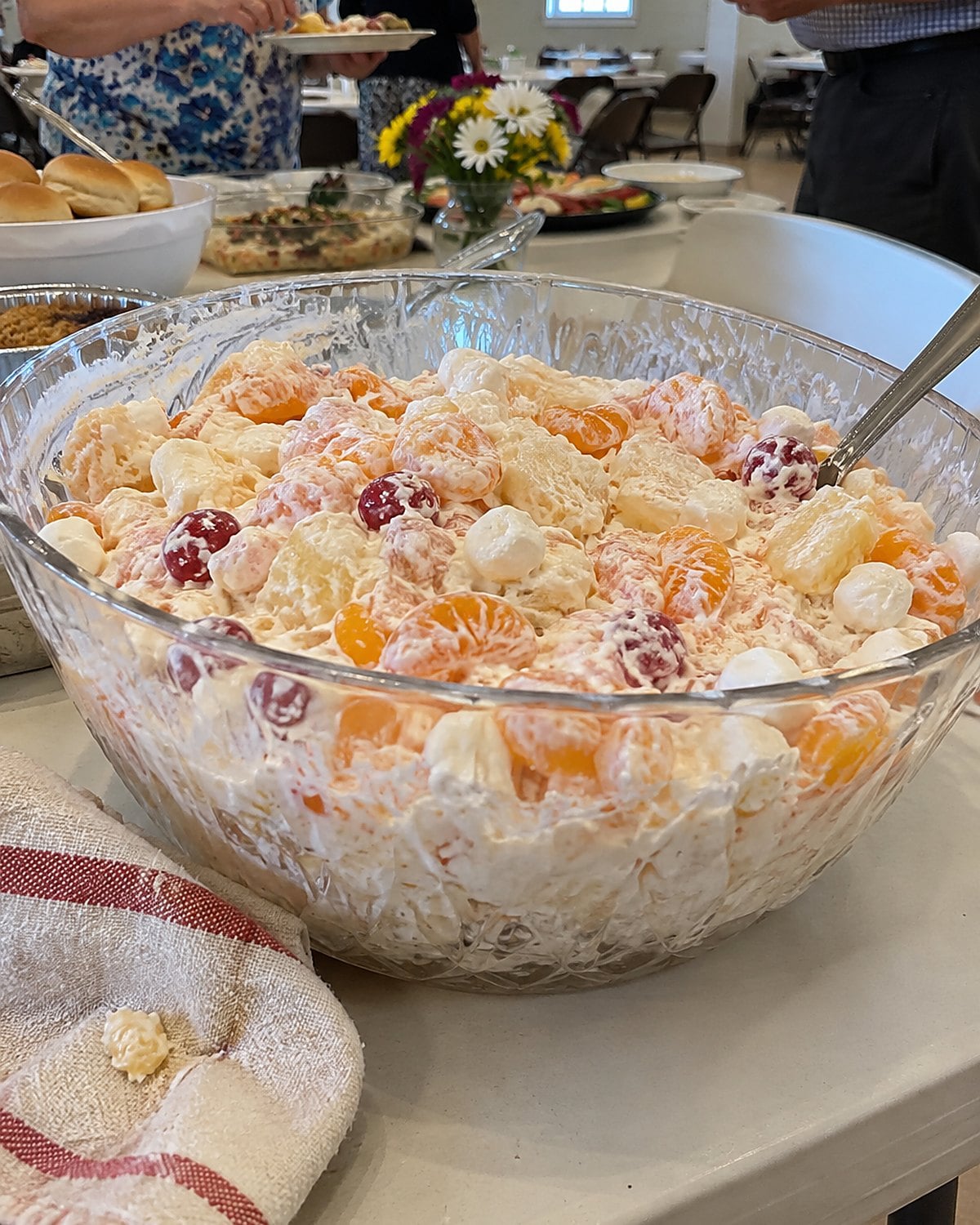 A large bowl of ambrosia with a serving spoon at the edge of a church hall table.