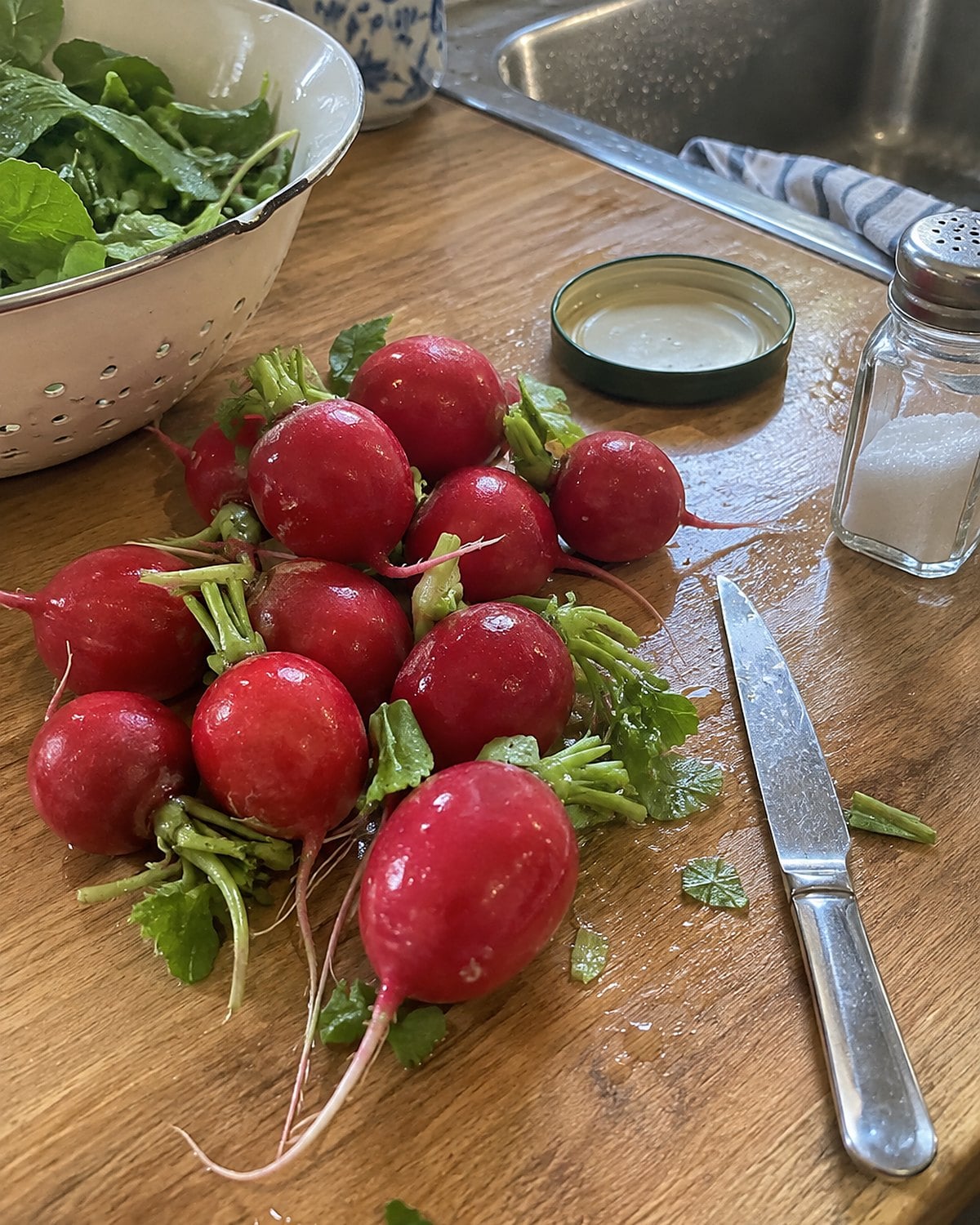 Washed radishes with trimmed leaves beside a butter knife and salt shaker on a counter.