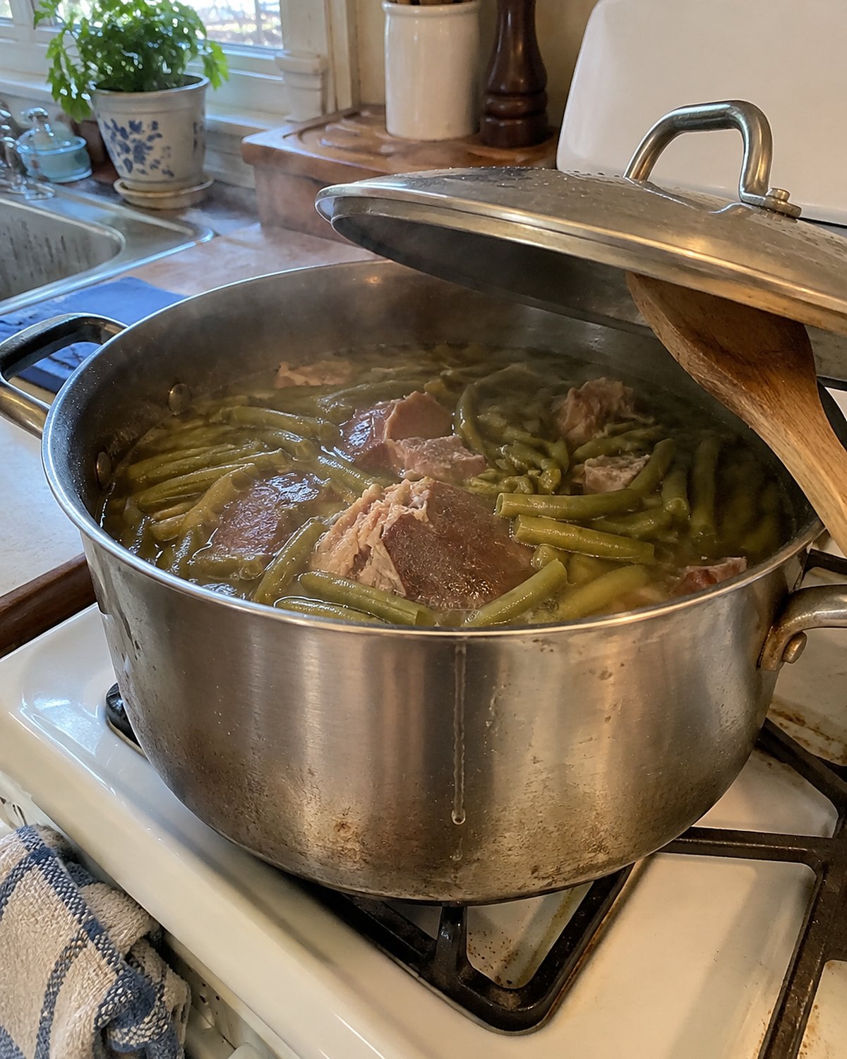A dented pot of green beans and ham hock on a white stove, its lid propped on a spoon.