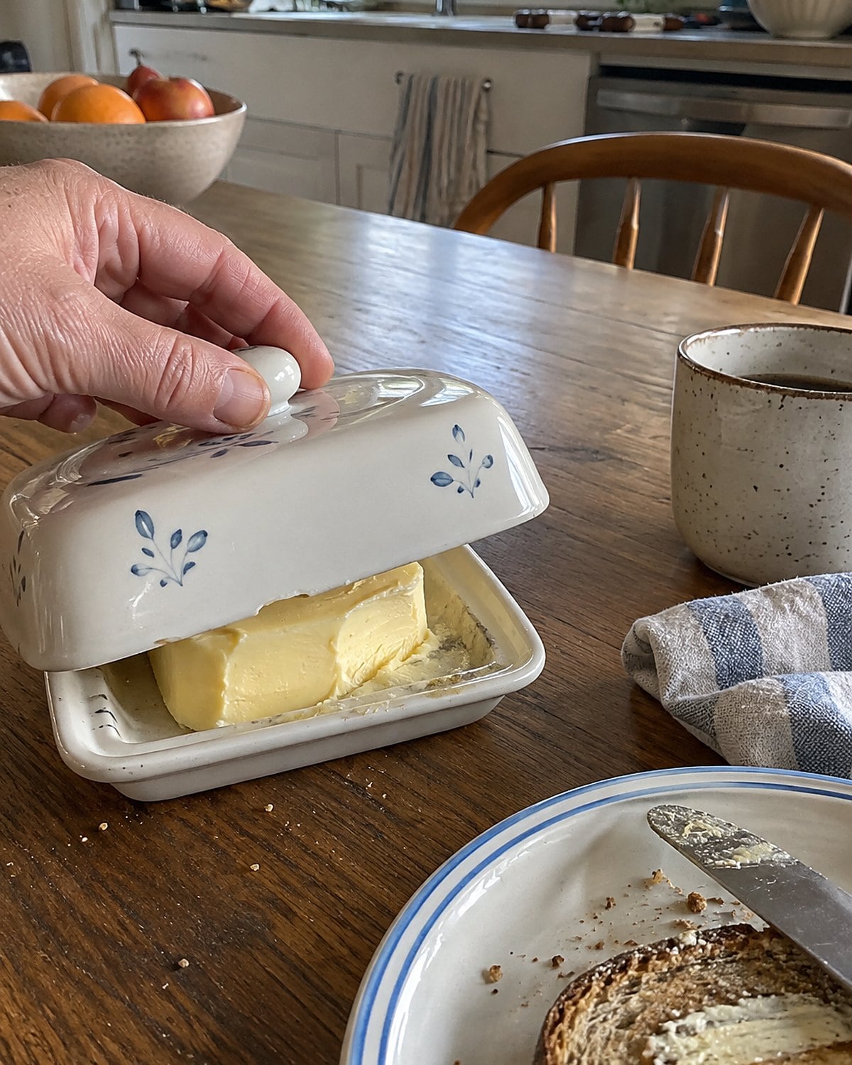 A covered butter dish with a few crumbs beside a dinner plate on a worn kitchen table.