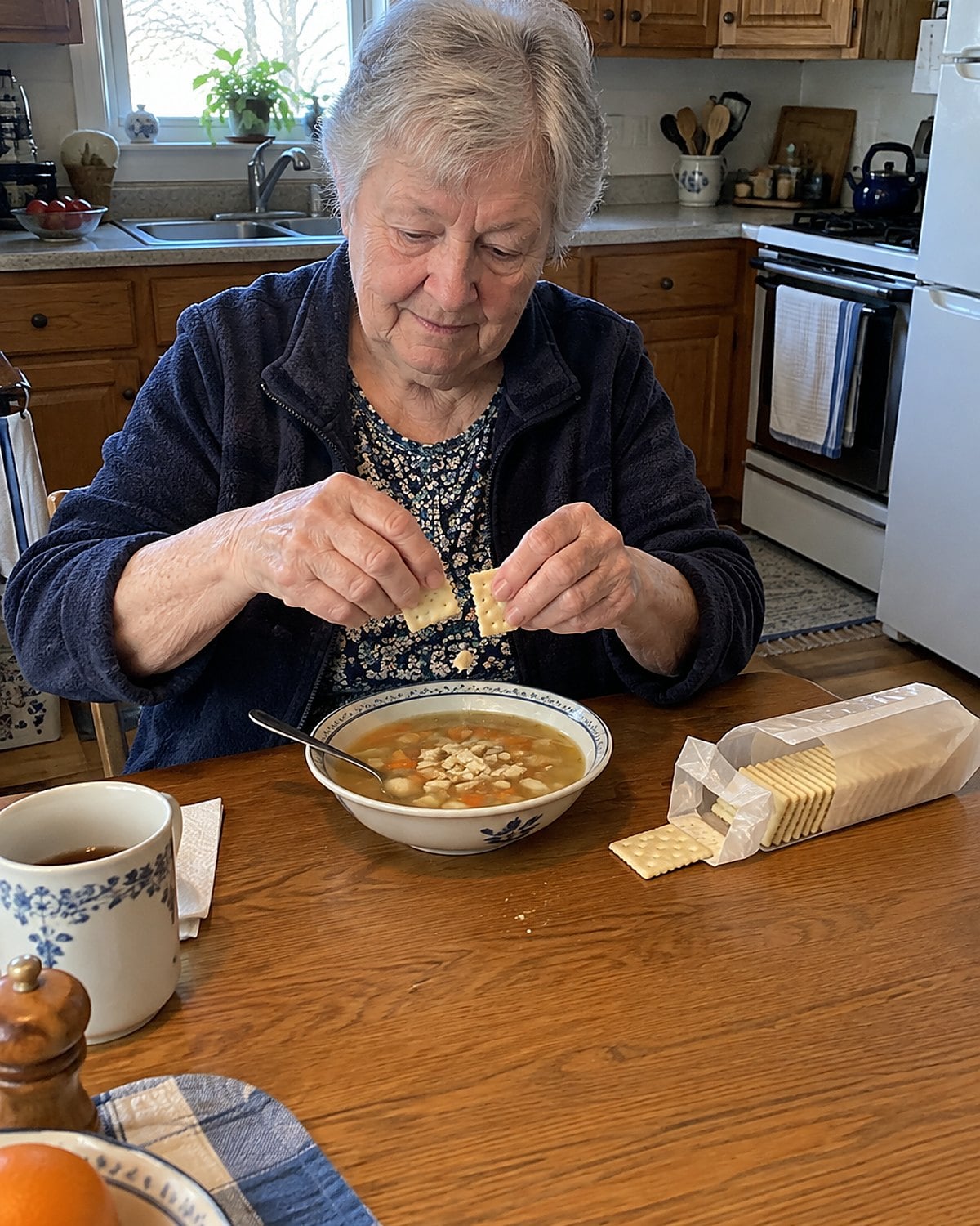 Hands crumbling saltines over soup while an opened cracker sleeve rests nearby.