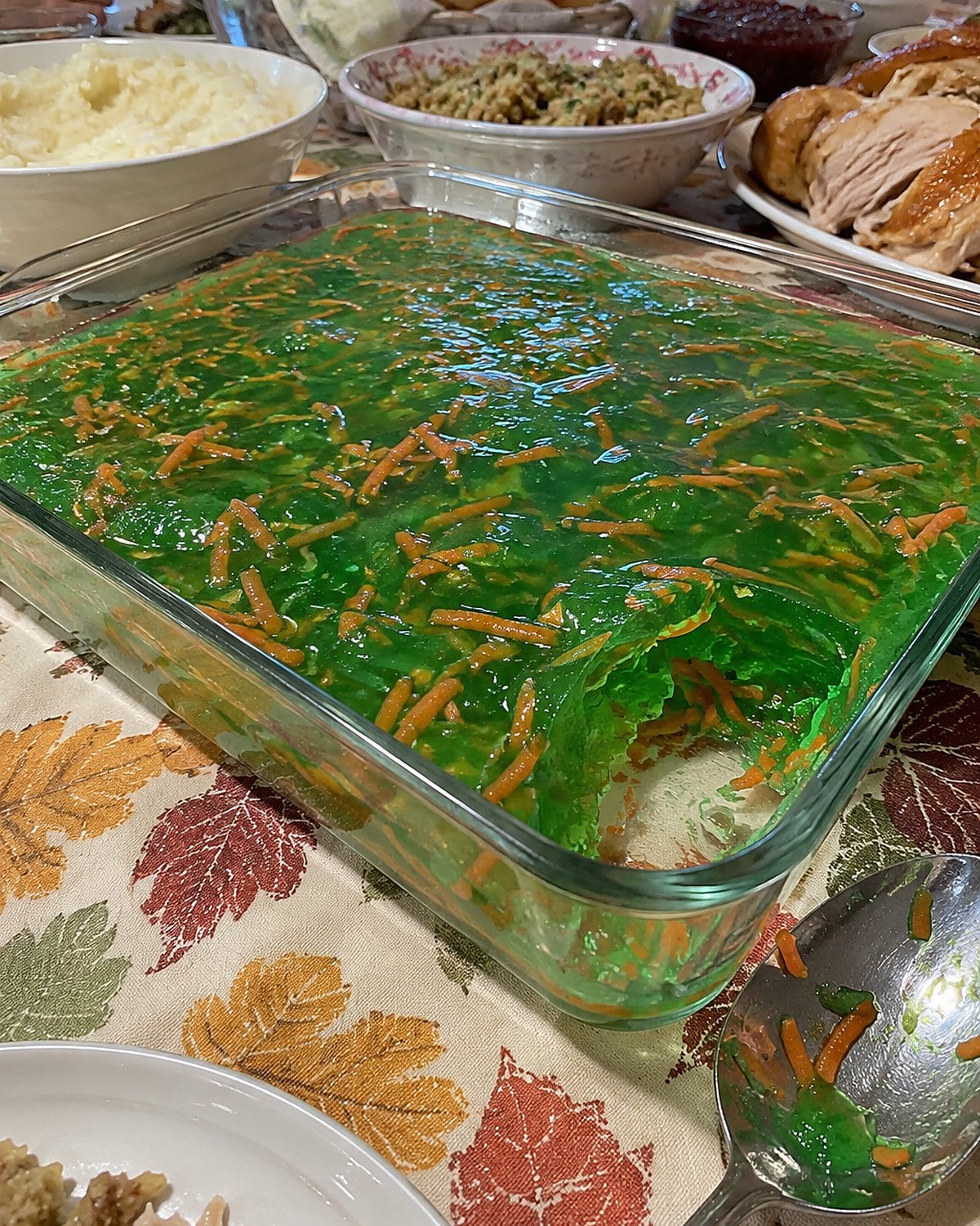 Green gelatin with visible carrot shreds in a glass dish on a crowded holiday table.