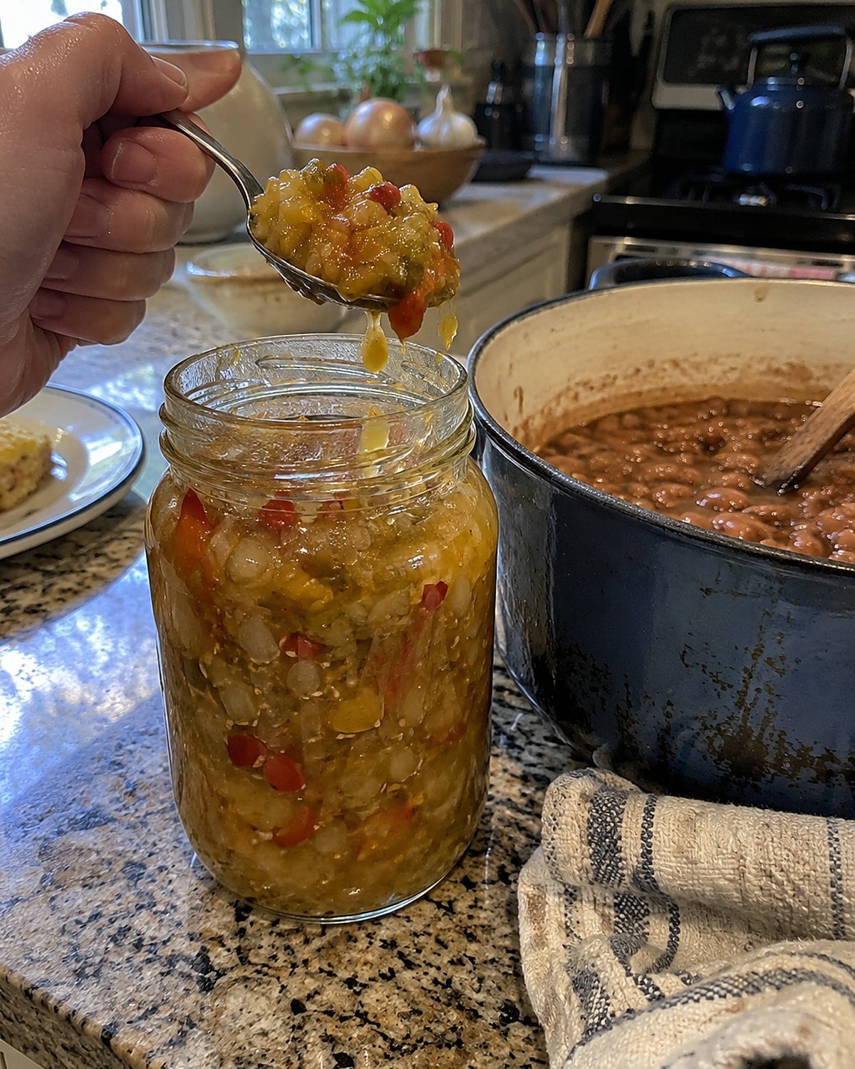 A spoon lifting chow-chow from an open jar beside a pot of beans.