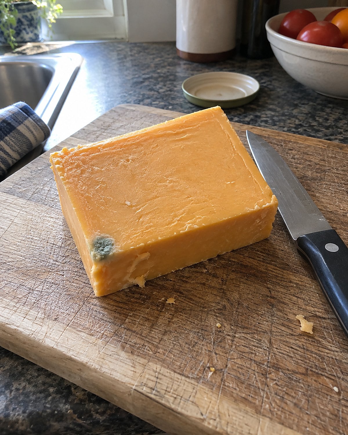 A block of cheddar with a small mold spot sits beside a clean knife on a wooden board.