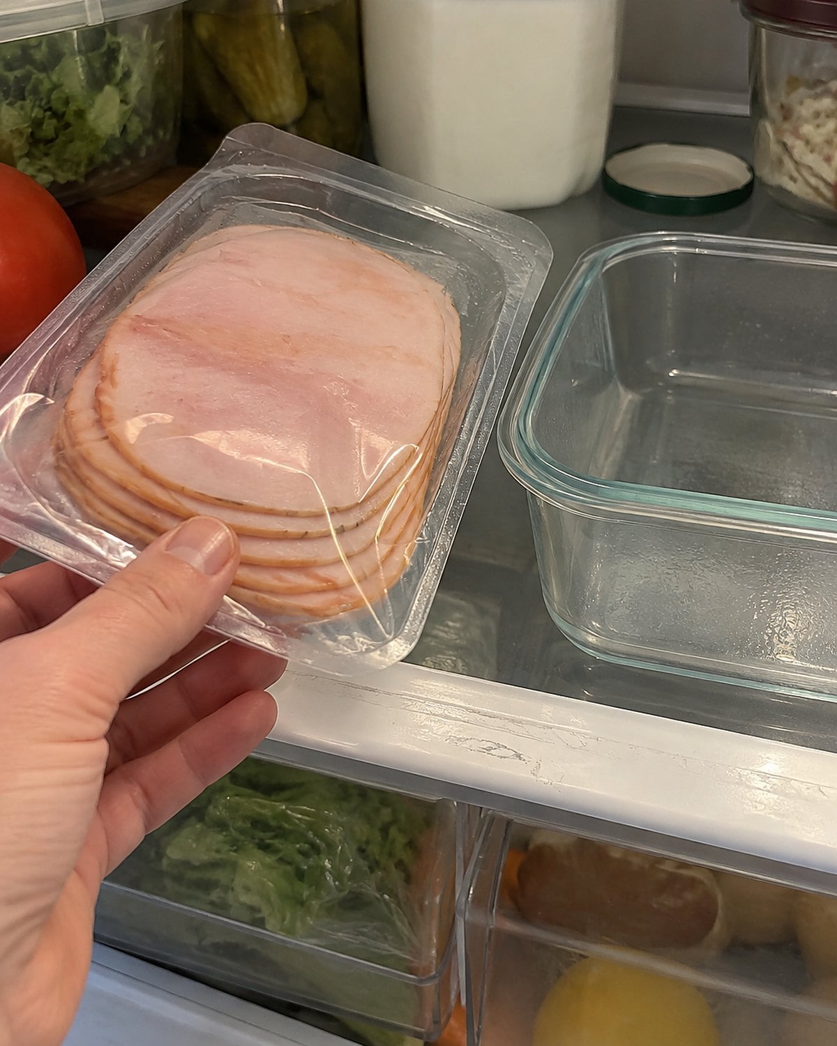 A sealed lunch meat package sits alone on a refrigerator shelf beside an empty sandwich container.