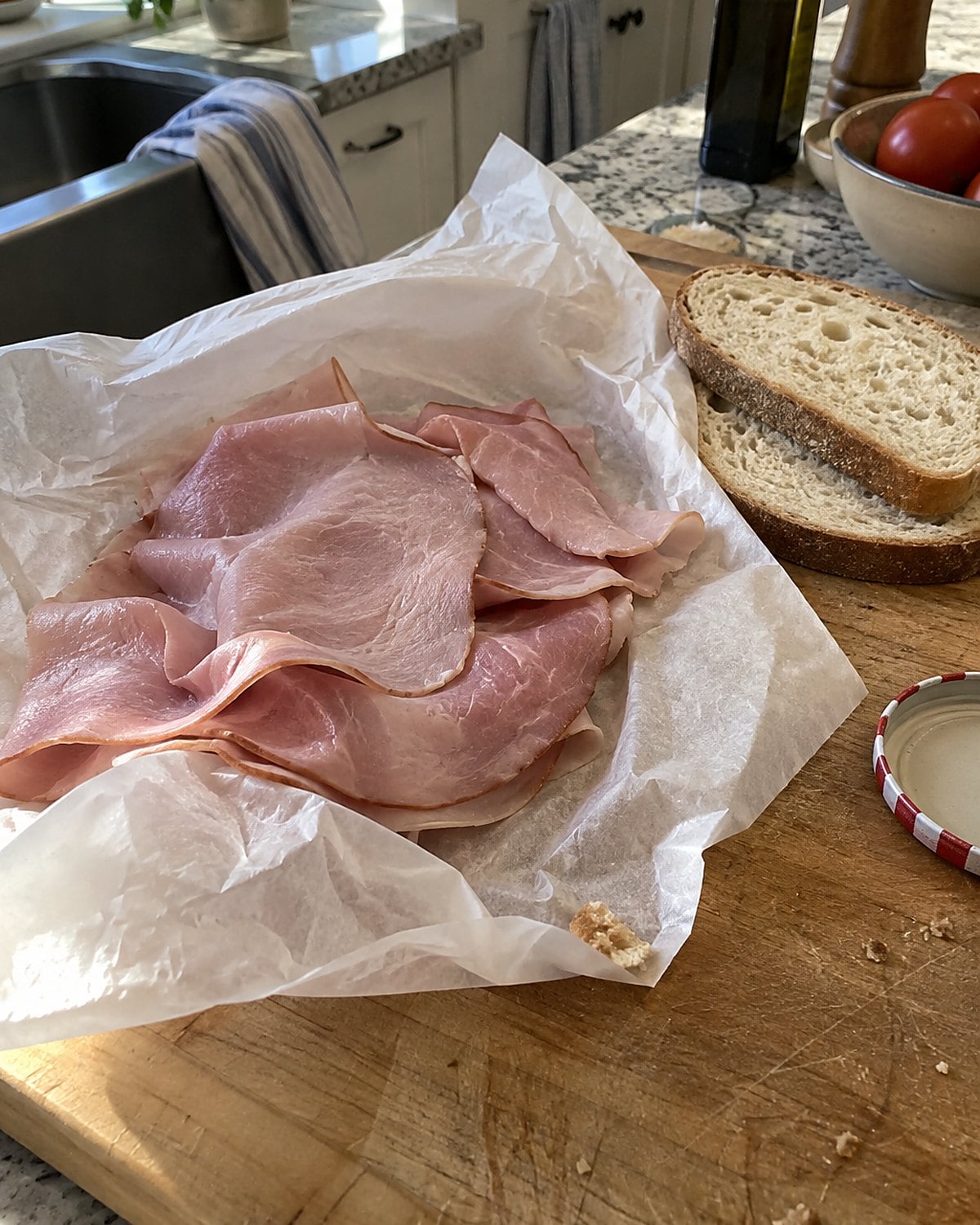 Deli ham wrapped in creased paper rests on a cutting board beside two slices of bread.