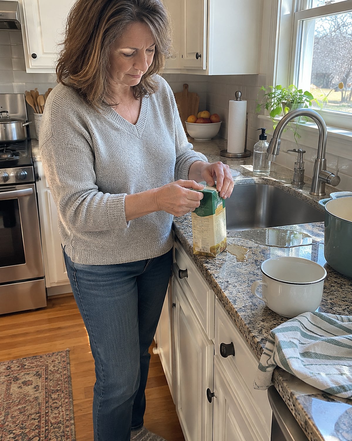 A hand folds the spout of a half-empty broth carton beside the kitchen sink.