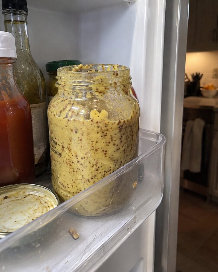 An old mustard jar stands among sticky condiment rings on a refrigerator door shelf.