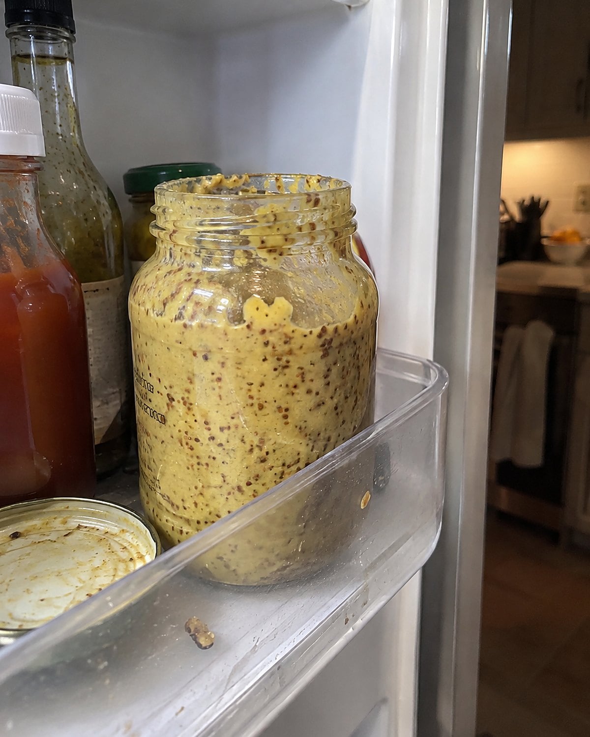 An old mustard jar stands among sticky condiment rings on a refrigerator door shelf.