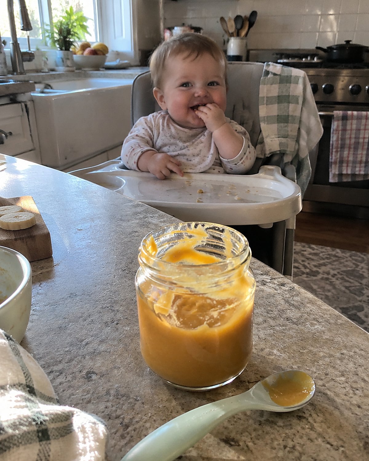 A spoon rests beside a partly used baby food jar on a kitchen counter near a high chair.
