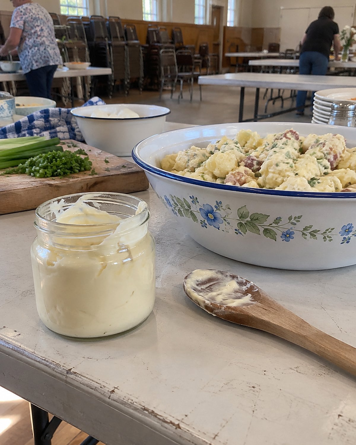 A small jar of homemade mayonnaise stands beside a bowl of potato salad on a church hall table.