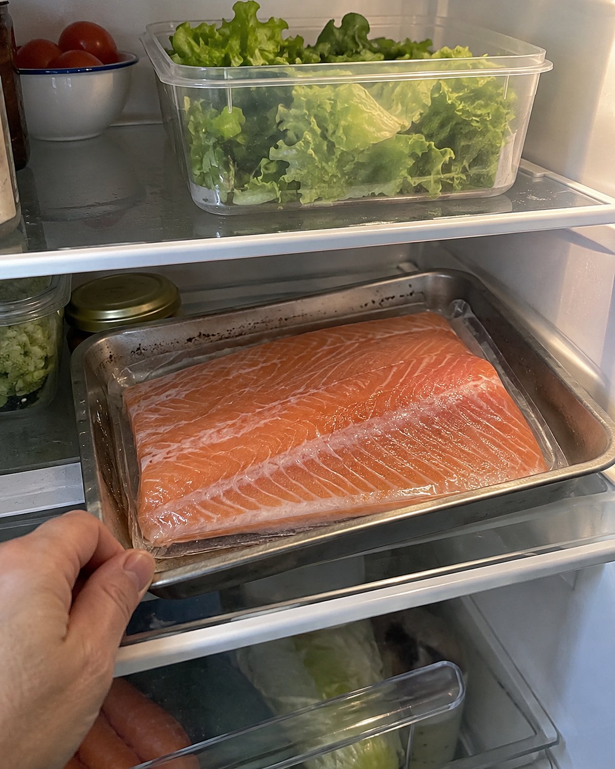A fish packet lies on a tray beneath a box of lettuce in the refrigerator.
