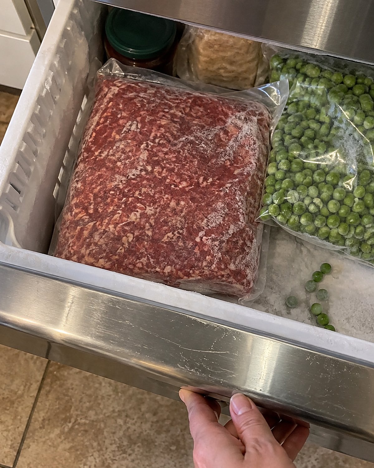 A package of ground beef rests beside a bag of peas in an open freezer drawer.