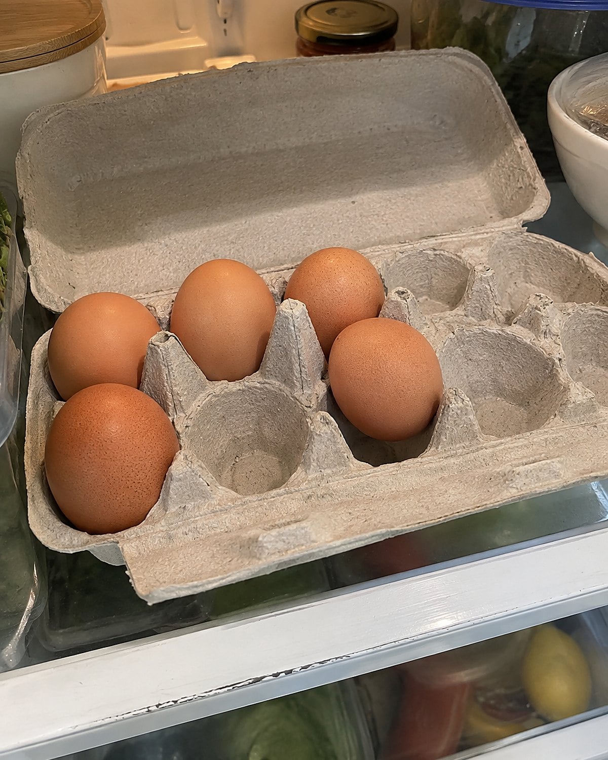 An open egg carton rests on an inside refrigerator shelf with several eggs missing.