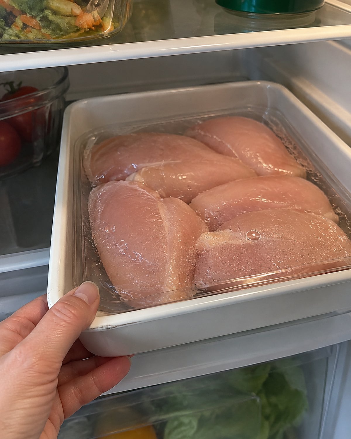 A sealed tray of raw chicken sits inside a rimmed container on the bottom fridge shelf.