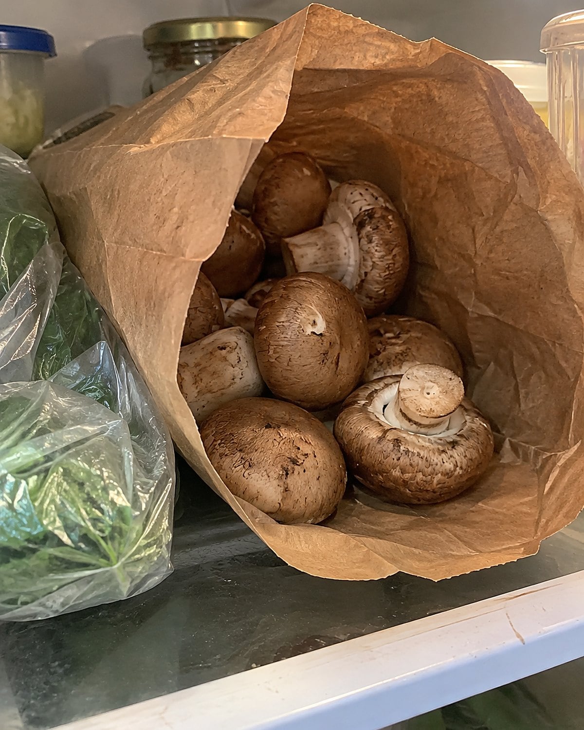 A brown paper bag holds loose mushrooms on a refrigerator shelf beside a folded plastic produce bag.
